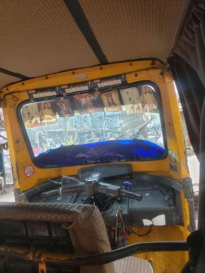This image captures the interior of a yellow auto-rickshaw, commonly known as a "Keke," in Alagbede, Nigeria. The vehicle's interior is somewhat cluttered. On the passenger side, a series of stickers or posters are affixed to the window, featuring religious imagery and text. Prominently visible are phrases like "My year of DOMINION" and "JESUS THE SAVIOR," alongside depictions of religious figures. The central window is severely cracked, with spiderweb-like fractures obscuring the view of the outside, suggesting damage.

The driver's area shows a steering mechanism, a handbrake, and some keys. A checkered cloth is draped over part of the dashboard. The seats appear to be covered with a woven material, possibly rattan or straw, on the passenger side and a darker, textured material on the driver's side. The overall impression is that of a functional vehicle that has sustained damage, likely to its windshield. The presence of religious iconography suggests a personal touch or a form of blessing for the vehicle. The setting appears to be urban or semi-urban, given the limited view of what might be a street or marketplace through the broken window. No specific time of day or weather conditions are evident.