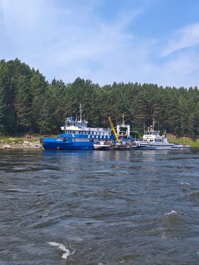 A blue and white vessel, identified by the inscription "ЕНИСЕЙ" (Yenisei) on its side, is docked on the right bank of a wide, turbulent river. Adjacent to it is a smaller, gray vessel. Both are situated in front of a dense, dark green coniferous forest that stretches across the horizon. The sky above is a clear blue with scattered white clouds. The river water is dark and shows signs of strong currents and ripples. The scene appears to be in the city of Porog, Russia, as suggested by the context.