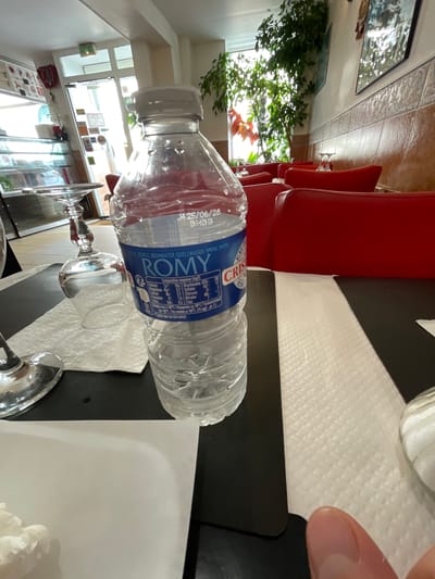 The image depicts an indoor scene, possibly a restaurant, with red seating booths visible. On a dark table, a plastic bottle of ROMY brand spring water is prominently featured. A partially visible wine glass sits to the left of the water bottle, placed on a white paper napkin. The background shows a window with abundant greenery, and to the left, a display case with items inside. The overall impression is of a casual dining setting, with the water bottle and glassware suggesting a meal is either in progress or about to begin. The label on the water bottle includes French text, indicating the origin of the product. The scene does not provide any explicit visual cues to pinpoint the exact location in Meaux, France, beyond the general impression of an establishment within that city.