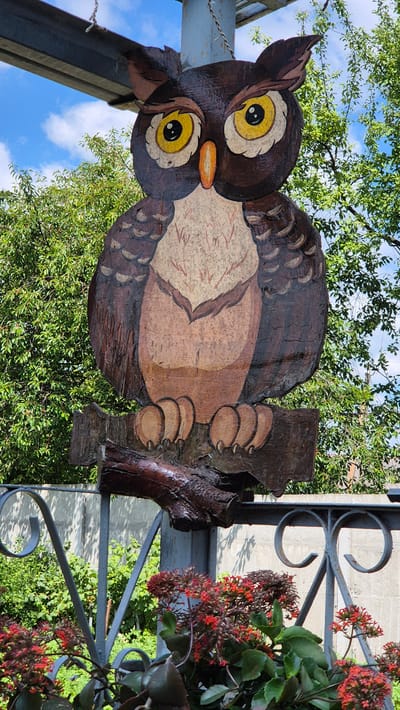 A carved wooden owl is displayed outdoors, suspended by chains from a metal pole. The owl is painted in shades of brown and cream, with large yellow eyes and an orange beak, perched on a wooden branch. The background features lush green trees and a bright blue sky with scattered clouds. In the foreground, a decorative metal fence with red flowers and green foliage is visible. The overall scene suggests an outdoor setting, possibly a garden or public space, in Цярэніцкі сельскі Савет, Belarus.
