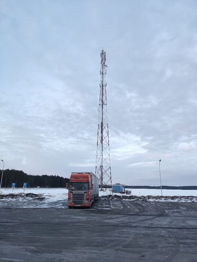An orange semi-truck sits parked on a snow-dusted asphalt lot. Behind the truck stands a towering red and white communications tower against a cloudy sky. Patches of snow cover the ground, and bare trees line the horizon in the background. A frozen body of water stretches out beyond the lot. A small blue structure and a lamppost are visible beside the tower. Parking signs stand near the trees.