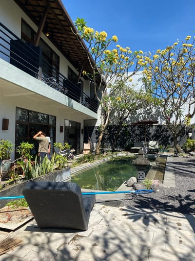 In Denpasar Barat, Indonesia, a sunlit courtyard features a rectangular pond with green water, bordered by dark grey stone tiles and gravel. Two lounge chairs with striped cushions are visible near the pond, partially shaded by an open dark umbrella. On the right side of the pond, a stone lantern and several rocks are present. The courtyard is surrounded by a building with a white facade and a wooden, sloped roof, with balconies on the upper floor. Lush green plants and trees with yellow flowers are scattered throughout the scene. In the foreground, a dark grey wicker lounge chair is positioned on a stone patio. A man wearing a green t-shirt and dark pants is standing near the building's entrance, with his hand on his head. The sky is clear and blue.