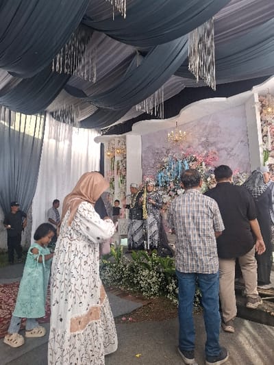 The image shows a wedding ceremony taking place in a tent, likely in Bojongsoang, Indonesia, based on the provided coordinates. The tent is decorated with draped fabric, creating a festive atmosphere. The ceremony is centered on a stage adorned with flowers, where a couple is standing, likely the bride and groom. Several guests are present, including a young girl in a blue dress, standing near a woman wearing a long white dress with floral patterns. Another woman is present, wearing a hijab and a long white dress with a floral pattern. In the foreground, a man in a plaid shirt stands with his back to the camera, while another man, dressed in a black T-shirt and beige pants, stands next to him. Both men appear to be observing the ceremony. The ground is covered with fallen leaves and a reddish-brown carpet. The overall scene suggests a joyful and celebratory atmosphere.