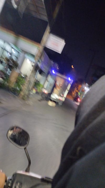 The image, taken at night in Bangsri, Indonesia, depicts a blurry scene from the perspective of a person riding a motorcycle. In the foreground, the handlebars and a rearview mirror of the motorcycle are visible. The mirror reflects a distorted image of the rider and the surroundings. The background features a street with a moving truck, its taillights illuminated. Buildings and vegetation are also faintly discernible along the roadside. The overall impression is one of motion and low light conditions, characteristic of a nighttime urban environment.