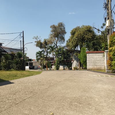 In Cibinong, Indonesia, a paved driveway curves towards a residential area under a clear blue sky. Green grass borders the left side of the driveway, leading to a house with a white exterior and a dark roof. Palm trees and other lush foliage are interspersed among the buildings. A light pole with numerous wires stands tall on the left, while another utility pole with electrical equipment is visible on the right. Further down the driveway, a wall constructed of rectangular stone blocks marks the entrance to a property. A small yellow leaf rests on the pavement.