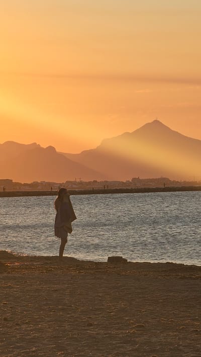 A lone figure stands on the sandy shore of Deveses, Spain, facing the water. The individual, silhouetted against a vibrant orange sky, appears to be holding something to their face, possibly a phone or camera. The sun is low in the sky, casting long shadows and illuminating the distant mountains with a golden hue. A body of water, likely the sea, stretches out before the shore, its surface rippled. In the background, a breakwater or pier is visible, with a few other distant figures walking along it. The overall scene evokes a sense of tranquility and the beauty of a sunset at the coast.