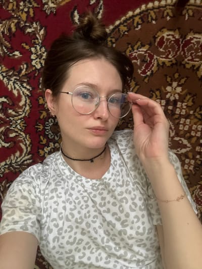 A female individual is depicted from an overhead angle, lying supine on a traditional red, beige, and brown patterned rug featuring floral and scrollwork designs. Her brown hair is styled in a messy bun. She wears large, round, silver-rimmed eyeglasses, and her left hand (viewer's right) is raised, with fingers lightly grasping the right side of the glasses frame. Her eyes are light, and her expression is neutral, directed at the camera. She is wearing a white short-sleeved t-shirt with a grey leopard-spot pattern. Her accessories include a black beaded choker adorned with a dark four-leaf clover charm and a lighter, possibly gold-colored, heart-shaped charm. Small, gold-colored, intricate earrings are visible in both earlobes. A thin, rose-gold colored bracelet with two small, interlocking circular charms is present on her right wrist (viewer's left). The scene is located in the city of Furmanov, Russia.