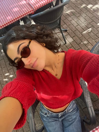 A young adult female is depicted in a high-angle selfie, seated outdoors on a patio in Orléans, France. She has dark, curly hair and wears tortoise-shell framed oval sunglasses, a red V-neck cropped knit sweater, and blue denim jeans. Her visible right ear features a gold stud earring, and she wears a double-layered gold necklace with small red beads and a heart-shaped pendant. The background shows dark, woven-style outdoor chairs and a table covered with a red and white checkered tablecloth. The ground is paved with light-colored rectangular bricks, illuminated by natural sunlight casting shadows.
