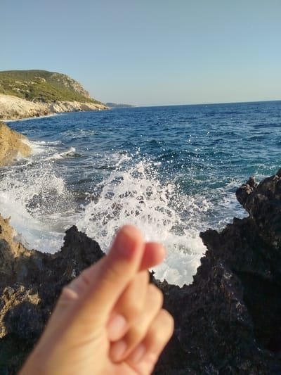 A hand forms a heart shape in the foreground, positioned in the lower-left quadrant of the image. Behind the hand, waves crash against dark, jagged rocks, creating a spray of white foam. The ocean extends to the horizon under a clear blue sky. In the background, a green, hilly coastline stretches along the left side of the frame, with some buildings visible in the distance. The scene appears to be a coastal landscape, likely in Karaburun, Turkey, given the vegetation and rocky shoreline.