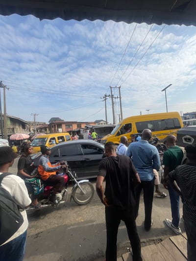 The image depicts a busy street scene in Akowonjo, Nigeria, under a bright, cloudy sky. A yellow minibus is prominent, likely a commercial vehicle. Several people are visible, some on foot, others on a motorcycle. A man in an orange and grey jacket rides a motorcycle, with a woman sitting behind him. Another motorcycle is parked nearby. People are dressed in casual attire. Buildings with awnings and signs line the street, suggesting a commercial area. Power lines and utility poles are also visible, indicating the presence of infrastructure. The road surface appears to be asphalt, with some oil stains. The overall impression is of a vibrant and active urban environment.