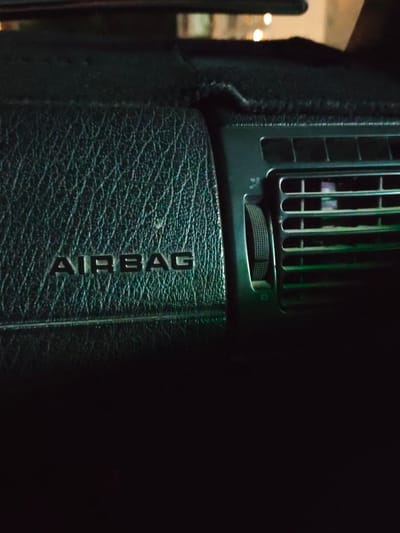 The image shows a close-up view of a car's interior, specifically the dashboard. The main subject is the textured dashboard material, which is a dark, possibly black or deep green, faux leather with a prominent cross-hatch pattern. Embossed into the dashboard on the left side is the word "AIRBAG" in silver, capitalized letters. To the right of the "AIRBAG" lettering, there is a car air vent. The vent has horizontal louvers and a rotary control knob on its left side for adjusting airflow. The overall lighting in the image is dim, suggesting it was taken at night or in a poorly lit environment. The background is dark and out of focus, with some faint, blurry lights that could be from streetlights or another vehicle. There are no people visible in the image. The scene is static, with no activity taking place. The focus is on the details of the car's dashboard components, highlighting the "AIRBAG" warning and the air vent. There are no explicit cues indicating the weather or a specific time of day beyond the dim lighting. The location context provided is Şardara, Kazakhstan, but this information is not visually evident from the image itself.