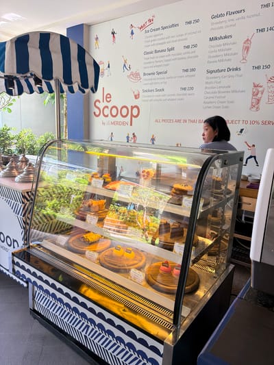 A bright, well-lit indoor scene depicts a dessert shop with a prominent display case filled with various pastries and cakes. The shop's name, "Le Scoop," is visible on the wall, alongside cheerful illustrations and menu items. The menu lists "Ice Cream Specialties," "Milkshakes," and "Signature Drinks," with prices indicated in Thai Baht.
In the foreground, a glass display case showcases an array of tempting desserts, including cakes, cookies, and tarts. The lighting in the case highlights the vibrant colors and textures of the treats. Behind the display case, a woman with dark hair and a striped shirt is looking to her right, seemingly attending to customers or overseeing the shop.
The environment appears clean and inviting, with decorative elements on the wall adding a playful touch. The overall impression is that of a bustling and appealing dessert establishment.