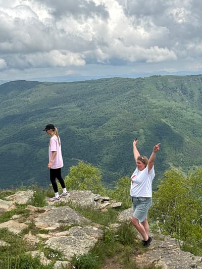 Two women are standing on a rocky cliff overlooking a lush green valley. The woman on the left, wearing a pink t-shirt, black leggings, a black baseball cap, and white sneakers, is looking down. The woman on the right, wearing a white t-shirt, denim shorts, and black sneakers, has her arms raised in a celebratory pose and is looking to her right. The background is a vast expanse of forested mountains under a cloudy sky. The location is identified as near Nizhnyaya Teberda, Russia.