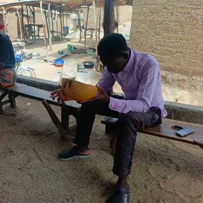 The scene is an outdoor setting, likely a communal or residential area, located in Boi B, Nigeria. In the foreground, a dark-skinned male, wearing a light purple long-sleeved shirt, dark trousers, and dark slip-on sandals, is seated on a light brown wooden bench. He holds a light brown, half-gourd-shaped object marked with "J1" or "F1" in both hands, looking down intently at it. To his right on the bench, a dark-colored smartphone with a silver back lies face-up. To the far left of the frame, partially visible, is the back of another person seated on the same bench, wearing patterned fabric. The ground is composed of sandy earth. The background features several rudimentary structures, including a prominent wall constructed of reddish-brown mud bricks to the right. The area appears to be partially covered by a makeshift shelter with a roof of branches or similar natural materials supported by wooden poles. Various objects are scattered on the sandy ground, including a blue plastic bowl, other containers, and dark pots. A blue and white motorcycle is partially visible to the left background.