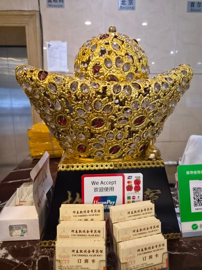 The image shows a decorative gold ingot with what appear to be glass or crystal embellishments, placed on a dark stand. In front of the ingot, there is a sign indicating "We Accept" with various payment method logos including UnionPay, VISA, and others. Several stacks of small, white cards with Chinese and Russian text are arranged in front of the stand. The text on these cards appears to be related to a hotel, possibly "Hunchun Kaijin Hotel" (珲春凯金帝宾馆). A green sign with a QR code is visible on the right side of the image, with Chinese text that translates to "Recommended to use". In the background, there are signs with city names in Chinese and English, including "Moscow", "Beijing", and "Seoul", suggesting an international context. The overall scene appears to be a reception area or lobby in Hunchun, China, likely at a hotel or similar establishment.