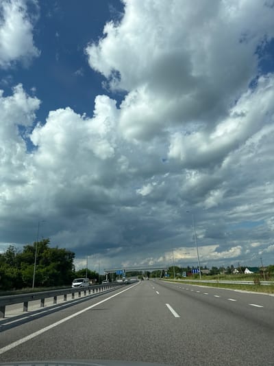 This is a landscape photograph, taken from a car driving on a highway. The orientation of the image is vertical, with the road occupying the right half and the sky the left. The road is asphalt with a white dashed line marking the lanes. A metal guardrail runs along the side of the road. On the left side of the road, there is a strip of grass, followed by a line of trees and some houses in the distance.

The sky is filled with dramatic, billowing clouds, some of which are dark and heavy, suggesting impending rain. Patches of bright blue sky are visible through breaks in the clouds. There are streetlights and road signs visible alongside the highway. One sign appears to be a blue directional sign. The time of day is likely daytime, possibly late afternoon, given the cloud formations and lighting. The weather appears to be changing, with a mix of sun and storm clouds.

The scene evokes a sense of travel and transition, perhaps on a journey through the Ukrainian countryside near Ksaverivka. The dynamic sky adds a sense of drama to the otherwise mundane highway scene. There is no visible text in the image, aside from what might be on the road signs.