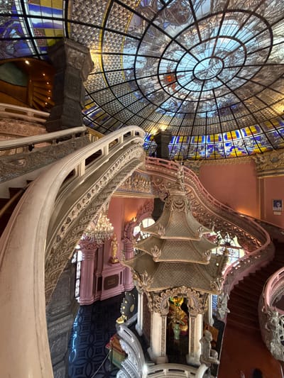 The image captures an ornate interior in the city of Samrong Nuea Subdistrict Municipality, Thailand. A massive stained-glass dome with intricate circular patterns dominates the scene, showcasing a blue and yellow color scheme. Adjacent to the dome is a winding, highly decorated staircase with white railings and detailed carvings in a light pink and beige color palette. The staircase ascends past openings offering glimpses of interior spaces and additional staircases. The environment exhibits a blend of architectural styles, showcasing Thai artistry.