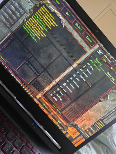 A black Acer laptop is positioned at an angle on a light-colored, wrinkled fabric surface, likely a bed or blanket, in Shotton, United Kingdom. The laptop screen displays the interface of the video game StarCraft: Remastered. The interface features a dark, industrial, metallic background with elements of pipes, grates, and machinery in grey, brown, orange, and yellow tones.
On the left side of the screen, a chat log displays messages in yellow and green text, repeating: "Connected to chat service.", "Welcome to StarCraft: Remastered!", "There are 78 games being played.", "There are 783 players online.", "Type /help for a list of commands.", "Joining default channel...", and "Joined channel Brood War En-1." To the right of the chat log, three red-outlined buttons with green text are visible: "Join", "Ranked", and "Ladder".
The right side of the screen displays a list of player names and channel information. At the top right, "kroks (aosfish)" is noted as "Online" with a green indicator. Below this, "Friends (0)" and "Brood War En-1 (45)" (indicating 45 participants) are displayed. A list of player names, each prefixed with an "SC" logo, includes "OnFire_", "Tantrum9", "colerbear", "ZergPractise", "exepm", "KingKeludis88", "noobbzone", "Samsonite01", "Kosto", "jjmayer", and "DustOfAngel", and "xSLEETX". Towards the bottom center of the screen, a partial "BLIZZARD ENTERTAINMENT" logo is visible on a textured metal panel. At the bottom right, three more red-outlined buttons with green text read "News", "Join Channel", and "Quit".
A portion of the laptop's black keyboard is visible in the lower left, featuring red-backlit keys. Visible keys include alphanumeric keys (e.g., "P"), function keys (e.g., "F10", "F12"), and navigation keys such as "PrtSc", "SysRq", "Pause", "Scr Lk", "Insert", "Delete", "Home", "End", "Page Up", "Page Down", and arrow keys. Two small blue LED indicator lights are illuminated above the keyboard, near the screen hinge. The overall lighting appears to be interior ambient light.