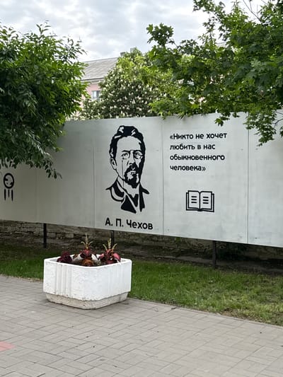 The image captures a street scene in Uryupinsk, Russia. A large, vertical white sign stands prominently on the street. The sign features a black and white portrait of Anton Chekhov, and beside his portrait, the name "A. П. Чехов" is written in Cyrillic. Above the portrait is a quote in Russian and an icon of a book. To the right of the sign is a white, concrete flower planter filled with red and green plants. The sign is surrounded by greenery, including trees. The paving stones of the sidewalk are visible on the right of the sign.