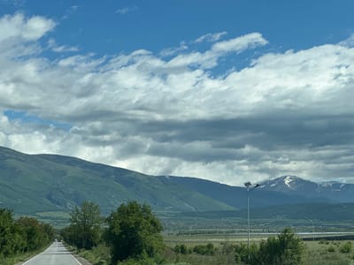 The image shows a road leading towards green, layered mountains under a cloudy sky. The road is asphalt, with white lines dividing the lanes. Trees and bushes line either side of the road in the foreground. In the middle ground, there is a plain with a village visible. A tall lamppost with a solar panel is standing in the vicinity. The mountains in the background have some snow cover. The photograph was taken in the vicinity of Marino Pole, Bulgaria.