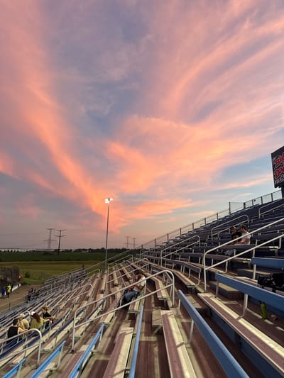 This photograph depicts a sunset scene at a stadium, likely in Joliet, Illinois, given the prominent presence of large electrical transmission towers in the background, which are characteristic of power infrastructure in the region. The sky is dramatically lit with streaks of pink and orange clouds, indicative of a vibrant sunset. In the foreground, tiered bleacher seating, typically found in sporting venues or outdoor event spaces, is visible. Some individuals are seated on the bleachers, observing the sky or engaging in conversation. The stadium lighting is on, casting shadows across the seating area. The overall environment suggests an outdoor event or a gathering taking place at dusk.