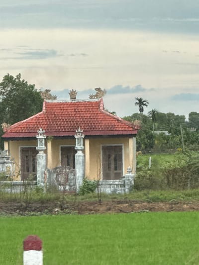 This is a medium-shot photograph of a small, yellow building with a red tiled roof. The building appears to be a temple or shrine, as indicated by decorative statues of mythical creatures on the roof ridge and ornate pillars flanking the entrance. The building has two prominent windows on its facade, and a decorative metal gate in front. The number "1079" is visible on a stone structure to the right of the gate. The building is situated in a rural setting, with lush green fields in the foreground and a dense backdrop of trees and vegetation. The sky is overcast with a few wispy clouds. A red and white marker post is visible in the lower left corner of the frame. The image is taken from a slightly elevated position, likely from a vehicle. The location is Hiếu Giang Commune, Vietnam.