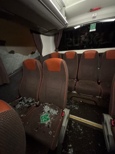 The image displays the interior of a passenger coach or bus, located in Hattencourt, France, exhibiting significant damage. A large section of a side window on the left is shattered, with numerous pieces of tempered glass scattered across several unoccupied brown fabric seats, which feature orange headrests, and onto the dark-colored floor of the vehicle's aisle. Overhead luggage compartments are visible above the seating area. Interior lighting is active, illuminating the scene, while the view through the intact windows reveals a dark exterior environment. A red emergency hammer is mounted near an intact window, which also bears a green emergency exit sticker. A green safety instruction sticker rests on one of the glass-covered seats. No individuals are present within the visible area of the vehicle.