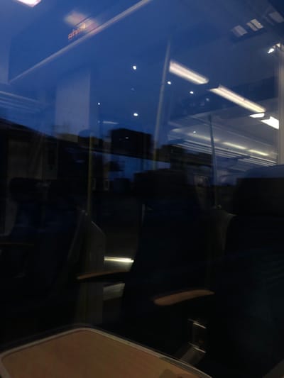 An abstract composition featuring reflections and interior elements within a train car. Reflected ceiling lights and building exteriors create a layered effect on the window surface. Dark seating and tables are visible within the train car's interior. The scene evokes a sense of movement and urban transit.