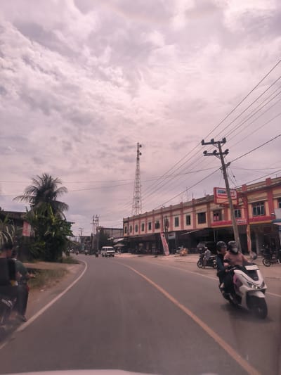 The image depicts a bustling street scene in Meukokuthang, Indonesia, under an overcast sky. A two-lane paved road, marked with a yellow dividing line, curves slightly to the left through the center of the frame.

In the foreground, several motorcycles are visible. On the right, two individuals wearing helmets ride a white scooter. On the left, a person on a black "Honda" motorcycle is partially seen. Further down the road, other motorcycles and a white van are in motion, indicating active traffic.

Along the right side of the road, a series of two-story commercial buildings or shophouses are prominent, featuring red and yellow exterior walls and multiple windows. A red and white "Alfamart" sign, identifying a convenience store, is clearly visible on one of these buildings. Utility poles carrying a dense network of overhead wires line the street, extending towards the horizon.

To the left of the road, tropical vegetation, including palm trees and dense green foliage, is abundant. In the mid-ground, a tall, metallic lattice communication tower (likely a cell tower) stands against the cloudy, somewhat pale sky. The overall scene suggests an ordinary daytime setting in a lively Indonesian town, characterized by daily commutes and local commerce under a heavily clouded sky.