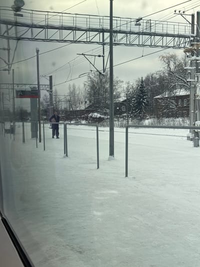 A snowy, possibly icy scene. The ground is covered in snow. Trees, some with snow on the branches, are visible. Overhead power lines and poles run across the landscape. A building is seen in the background. A person stands in the snowy expanse, possibly at a train station or platform. The image appears to be taken from inside a vehicle, possibly a train or car, due to the reflected interior.