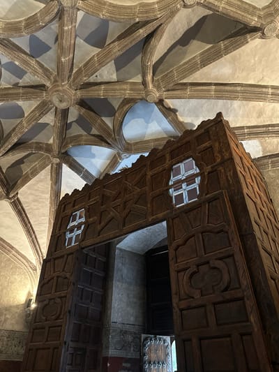 The image shows the interior of a colonial-era building, likely a church or monastery, in Huejotzingo, Mexico. The most prominent feature is a massive, intricately carved wooden door structure, appearing to be a confessional or a similar partition. This structure is made of dark wood with geometric patterns and ornate carvings. Above the doorway, there are two small, stained-glass windows with a diamond pattern. The ceiling is a striking example of Gothic architecture, with ribbed vaults and stone embellishments, suggesting a significant historical and artistic value to the location. The walls have some faded murals or decorative painting towards the bottom. The lighting is dim, with some light sources casting shadows, enhancing the dramatic effect of the architecture and the imposing wooden structure. No people are visible in the frame.