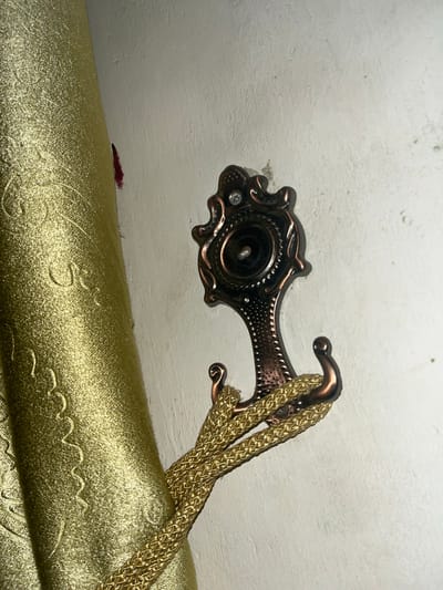 The image shows a decorative bronze curtain tie-back hook mounted on a white wall. The hook has an ornate design and is holding a gold braided cord, which is part of a curtain. The curtain itself is made of a textured gold fabric with a raised pattern. To the left of the hook, a small red smudge is visible on the wall. The scene is indoors, likely in a residential setting. This scene is not located in the city of Lambaro Kueh, Indonesia.