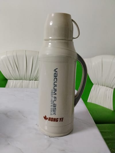 A light beige vacuum flask with a grey handle stands upright on a white marble patterned table. The flask has text printed on it that reads "VACUUM FLASK KEEP HOT & COLD" vertically, with a small red maple leaf symbol above the word "HONG YE" in red letters. The flask is positioned in the center of the frame, with the back of two green and white tufted sofas visible behind it. The environment appears to be indoors, possibly a living room. No people are present in the image. The location is Almaluu, Kyrgyzstan.