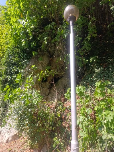 The image shows a street lamp with a spherical, translucent top, positioned in front of a rocky, overgrown embankment. Lush green foliage, including ivy and deciduous trees, covers parts of the embankment and hangs over the rocks. The lighting suggests it is daytime, with sunlight filtering through the leaves, creating dappled shadows. The street lamp is a tall, silver pole with a visible joint and a silver base. The embankment itself appears to be a natural rock formation with some soil and debris at its base. There is a dark opening or cavity visible within the rock face. The overall scene conveys a natural, somewhat wild environment integrated with urban infrastructure. This scene is likely located near the entrance to the Teplice nad Bečvou spas, a well-known tourist destination in the Czech Republic.