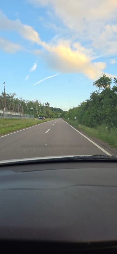 The image shows a view from inside a vehicle driving on a road in Wallerfangen, Germany. The road ahead is paved and has white lane markings. On the left side of the road, there is a fence, some trees, and what appears to be a parking lot with a few dark-colored cars. A light pole is visible near the fence. On the right side of the road, there are grassy verges and dense green trees and bushes. The sky is blue with scattered white and yellow clouds, and a contrail is visible. The lower portion of the image shows the dashboard of the vehicle, out of focus.