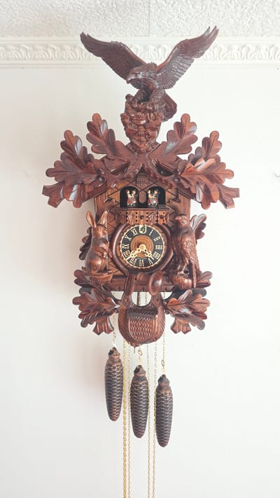 The image shows a detailed, carved wooden cuckoo clock hanging on a white wall. The clock features an eagle at the top, an ornate design with oak leaves surrounding the clock face, a rabbit on the left, and another bird, likely a raptor, on the right. Two small figures are visible in an upper window, possibly representing cuckoos. Below the clock face, there is a small horn and a decorative basket-like element. Three pinecone-shaped weights hang from gold chains below the main body of the clock. The wall behind the clock has a white textured pattern at the top, suggesting it is either wallpaper or a textured ceiling. The clock is intricately carved, with a dark brown wood finish. There is no discernible information in the image that places it in Crailsheim, Germany; the description is limited to the visual content of the image itself.