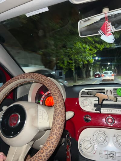 The scene depicts the interior of a red Fiat 500 automobile at night, looking forward through the windshield onto a street in Lomas de Zamora, Argentina. The steering wheel, partially obscured by a visible hand, is covered with a leopard-print fabric cover. The dashboard features a prominent red panel across its upper section, contrasted with white lower controls for climate and media. The instrument cluster is illuminated with an orange glow, displaying a speedometer. A silver and black phone holder is clipped to the central air vent. Keys with a red fob hang from the ignition switch. A miniature red high-top sneaker air freshener hangs from the rear-view mirror.
Through the windshield, a tree-lined street is visible, illuminated by distant streetlights. The environment shows signs of motion blur, indicating the vehicle is in transit. Ahead, two cars are visible in the distance: a red sedan followed by a white sedan. The road surface appears to be asphalt. The surrounding area consists of residential or urban development, obscured by darkness and motion.