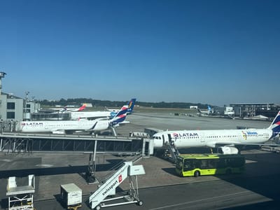 The image captures an airport tarmac scene at Guarulhos, Brazil, under a clear, bright blue sky with faint distant contrails. Viewed from an elevated position within a terminal building, the foreground features a large white LATAM Airbus A320 family aircraft (registration PT-MIE) parked at a gate. The plane displays "LATAM BRASIL" text and a colorful "BRASIL" logo on its fuselage. It is connected to a passenger boarding bridge at its forward door, and a separate set of mobile stairs is positioned at a rear door. A lime green "GRU AIRPORT" bus is parked alongside the aircraft, with a driver visible in its cab.

In the mid-ground, to the left, another white LATAM aircraft (registration CC-MEH) is also parked at a gate, connected to a second passenger boarding bridge. A white Swissport mobile stair (SWB10363) is visible near this aircraft. Further back, partial views of additional LATAM aircraft tails, an Iberia tail (red and yellow stripes), and other indistinct aircraft are visible. Various ground support equipment, including dnata-branded baggage loaders, PROTAIR containers, and other dollies, are scattered across the concrete tarmac. Airport buildings and a distant treeline frame the horizon. A few individuals are discernible on the mobile stairs of the foreground aircraft, suggesting passenger activity.