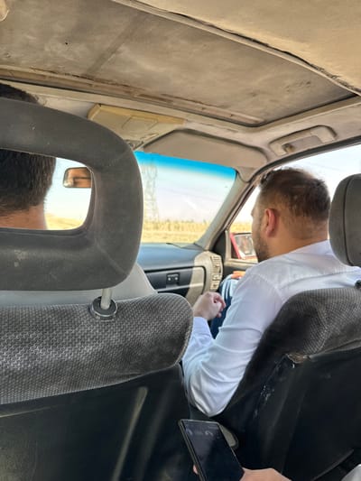 The image depicts the interior of an aged vehicle, viewed from the rear passenger seat looking forward, within the City of Adra labor, Syria. Three individuals are present: the photographer, whose hand is visible in the bottom right holding a black smartphone with a dark screen; a front passenger seated in the right front seat; and a driver, partially obscured by the front passenger's headrest.
The front passenger is a male with short, dark brown hair and a visible beard, wearing a white long-sleeved collared shirt and blue jeans. He is positioned facing forward, slightly turned to his right, with his hands clasped in his lap. The driver is also male, with short dark hair, largely obscured by the front passenger headrest and the perspective.
The car's interior shows signs of wear. The light-colored headliner above is visibly sagging in multiple areas and has some discoloration. A sunroof panel is present but closed. The front passenger seat is upholstered in a dark gray patterned fabric, with the headrest in a matching darker gray. The back of the seat shows some darker, possibly worn or stained, patches. The dashboard is dark, featuring air vents and control panels. A portion of the front passenger door panel, appearing red, is visible.
Through the windshield, a clear, sunny exterior environment is visible. A light-colored road extends into the distance, flanked by dry, light brown fields or open land. A large metallic power transmission pylon is visible in the mid-ground on the left side of the road. The sky is bright blue, fading to a lighter hue at the horizon.