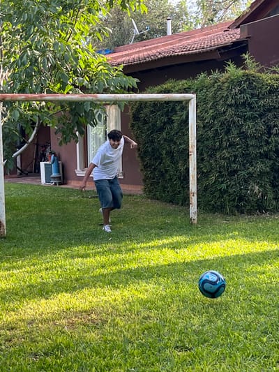 A young man in a white t-shirt and denim shorts points towards a blue soccer ball in mid-air. He stands on a green lawn near a weathered white metal goalpost. Behind him, a building with a brown roof and windows is partially obscured by lush green foliage. Sunlight casts shadows on the grass, suggesting a bright, sunny day.