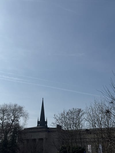 This image depicts an outdoor daytime scene in Bad Nauheim, Germany, featuring the Dankeskirche (Protestant Church) and surrounding environment. The upper two-thirds of the frame are occupied by a clear, light blue sky with faint, linear contrails visible in the upper mid-section.
The lower third of the image is dominated by architectural structures and bare deciduous trees. A dark, slender spire, characteristic of the Dankeskirche, with a sharply pointed apex and smaller decorative elements along its lower shaft, rises prominently against the sky. This spire originates from a large, light-colored stone building exhibiting classical architectural elements, including columns and an inscribed frieze on its facade. The building's flat roof is visible around the spire's base.
Bare deciduous trees with visible branch networks frame the structures, obscuring parts of the buildings on the left and right sides of the image. Their lack of foliage indicates a dormant season. To the right of the main church building, another section of a light-colored structure is partially visible, from which a white banner or sign with indistinct red markings hangs, partially obscured by tree branches. No individuals, vehicles, or active interactions are depicted within the scene. The lighting suggests ambient daylight, with no harsh shadows visible.