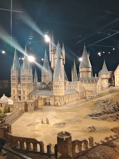 The image depicts a detailed model of Hogwarts Castle, the fictional school of witchcraft and wizardry from the Harry Potter series. The model showcases the castle's towers, courtyards, and surrounding landscape, including the greenhouse. The model is situated indoors, likely in a display setting, with spotlights illuminating the scene. The lighting suggests the setting might be the Warner Bros. Studio Tour in Watford, United Kingdom, where a large-scale model of Hogwarts is a primary attraction. No people are visible.