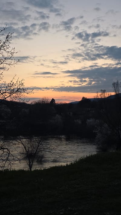 The image depicts a landscape scene near Byala Reka, Bulgaria at sunset. The sky is a mix of light blue and grey clouds, transitioning to orange and pink hues near the horizon. The silhouette of a mountain range is visible in the background. A body of water reflects the sky's colors and is lined with trees and vegetation. Bare tree branches are in the foreground. The vegetation on the right side of the frame is dark green, indicating a landmass. The overall scene suggests a rural or natural setting.