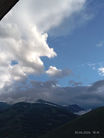 This media file depicts a scenic mountainous landscape, likely in Tyrnyauz, Russia. The scene is dominated by a dramatic sky filled with a mix of bright white and dark, stormy clouds. The sun appears to be setting or has recently set, as indicated by the contrasting light on the clouds and the subdued lighting of the mountains. The mountains themselves are covered in lush green vegetation and shrouded in mist, particularly in the valleys and on the higher peaks, suggesting a humid or post-rainy atmosphere. The presence of power lines and pylons running up the mountainsides suggests human infrastructure within this natural setting. The overall mood is one of serene, almost majestic, natural beauty, with a hint of the dramatic due to the dynamic cloud formations and rugged terrain.
Visible text in the image reads "30.06.2026 18:41", indicating the date and time the photograph was taken. This provides a specific temporal context to the image, placing it on the evening of June 30, 2026. There are no people visible in the image.