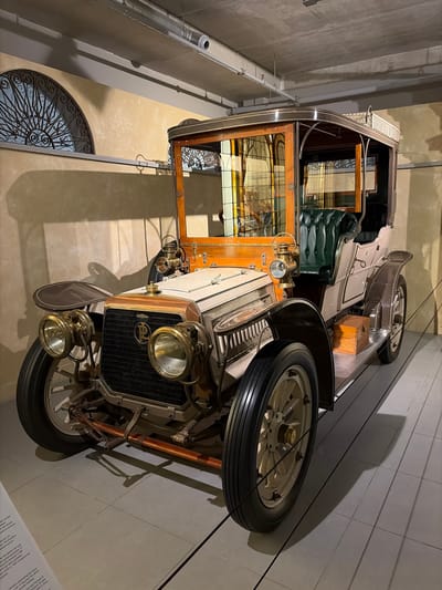 This image, rotated 90 degrees clockwise, displays an antique automobile, likely in a museum setting in Wassenaar, Netherlands. The car is largely cream or light beige with dark brown or black accents and features polished wooden elements.
The front of the car is visible, showcasing large, round, brass or gold-colored headlights and a prominent dark radiator grille with a logo that appears to be a stylized "PL" or similar design. Below the headlights, the front wheels with cream-colored spokes and black tires are visible. A spare tire is mounted on the side, near the front passenger area.
The interior of the car is partially visible, featuring a tufted, dark green leather seat (possibly driver's side), and ornate wooden paneling around the windows. The windows themselves have decorative, possibly stained glass or etched patterns. A rear view of the car shows a mesh luggage rack and another spare tire mounted on the side. The overall design suggests a luxury vehicle from the early 20th century.
The setting appears to be an indoor exhibition space with plain walls. Strong shadows are cast on the walls by the car and its components, suggesting artificial lighting. In the background, an ornate, semicircular metal grate or window treatment is visible.
Visible text from a display plaque, partially translated from Dutch and English, mentions "Panhard & Levassor," indicating the car's manufacturer. Phrases like "vierversnel" (four-speed), "autoproducenten" (car manufacturers), and "wordt in 1965 overgenomen" (was taken over in 1965) provide historical context about the company. Other fragments like "partition separating the driver" and "detachable hardtop" describe specific features of such vehicles.