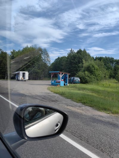 A roadside view from a vehicle shows a white kiosk with an open serving window and a person standing near it. To the right, a small blue and red open-air stall displays various items. A green minivan is parked in a grassy area behind the stall. The scene is set against a backdrop of dense green trees under a partly cloudy blue sky. The foreground shows the asphalt road with a white lane marking and a car's side mirror reflecting the sky and trees. The specific location within Ilyanskiy selskiy Sovet, Belarus, is a rural roadside setting.