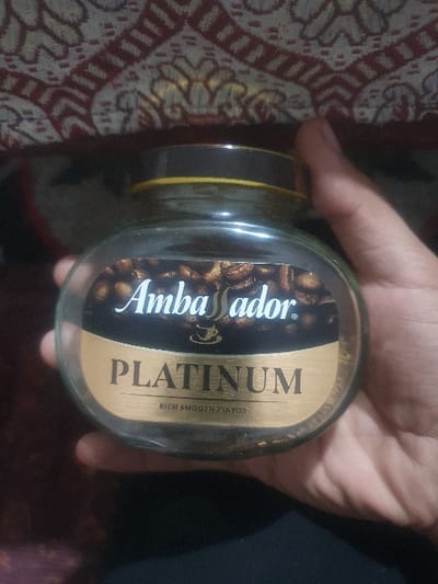 A person is holding a jar of "Ambassador Platinum" instant coffee. The label features coffee beans and the words "Rich Smooth Flavor." The jar has a yellow and brown lid. The background is a mix of patterns, with a reddish-brown patterned fabric on the left and a dark, possibly black, material on the right. The hand holding the jar is slightly out of focus.