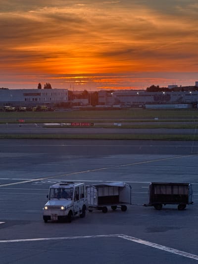 This is a vertical, medium-shot photograph of an airport tarmac at sunrise, featuring a tow tractor pulling two cargo trailers. The sky is a vibrant orange and yellow, with wispy clouds streaked across it. In the background, a long, low-rise industrial building with the "SAFRAN" logo is visible. The tarmac is a dark gray asphalt with white and yellow lines indicating taxiways and runways.
In the foreground, a white tow tractor, identified by "AZ081" on its windshield, is moving from left to right. It has its headlights on, illuminating the asphalt ahead. The tractor is pulling two trailers. The first trailer is a gray, open-top cart with a white tarp covering its contents. The second trailer is a darker, enclosed cargo container with a metal grate on its sides and a closed door. Both trailers have visible wheels and appear to be laden with goods. There are two individuals visible inside the cab of the tow tractor, facing forward.
The location is the Brussels Airport in Zaventem, Belgium. The scene captures a moment of activity on the tarmac during the early hours of the morning, under a dramatic sunrise sky.