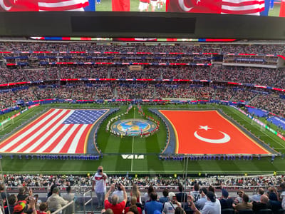 This is a high-angle, wide shot of a soccer stadium during a pre-game ceremony. The field features two massive flags, one of the United States and one of Turkey, spread out across the turf. Between the flags, a circular graphic displays the Earth with "FIFA WORLD CUP 2026" text. A line of players in blue uniforms stands on either side of the field, flanking the flags and the central graphic.
The stadium seating is filled with a large crowd of spectators, many wearing red, white, and blue. Banners with "LOS ANGELES" and "FIFA WORLD CUP 2026" are visible above the stands. A large digital screen is mounted at the top of the stadium, displaying the flags of the United States and Turkey.
In the foreground, a man in a white shirt with a Turkish flag emblem is taking a photo with his phone. Other spectators are also holding up their phones to capture the event. The scene is set at SoFi Stadium in Inglewood, California, a venue for the 2026 FIFA World Cup.