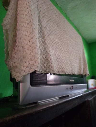 A television set is partially covered by a cream-colored, textured fabric. The television appears to be a flat-screen model with a silver bezel and a black speaker grille. The brand name "ASATRON" is visible on the front of the television. The television is positioned on a dark wooden surface, likely a table or shelf, in front of a bright green wall. The fabric cover has a repeating pattern of interconnected shapes. The lighting in the image is somewhat dim, casting shadows. The overall scene suggests an indoor setting, potentially a home. The context provided indicates this location is in Sukamanah, Indonesia.